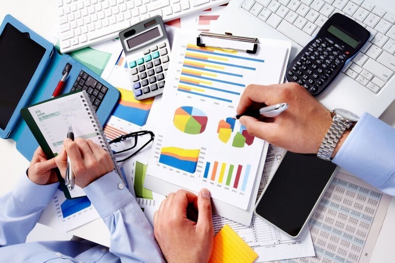 Imagem mostra várias duas pessoas analisando alguns relatórios financeiros, com celular e calculadora.