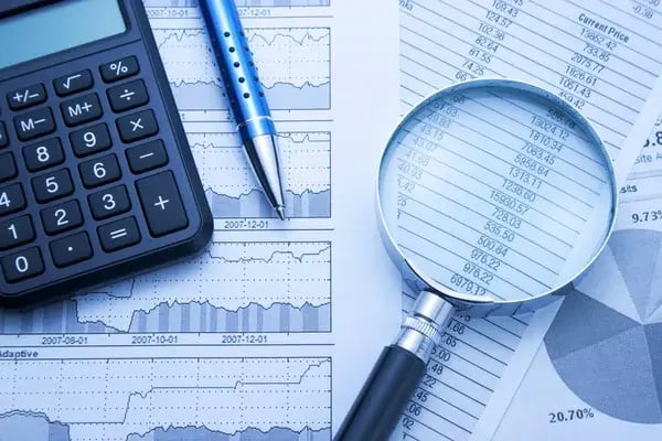 A imagem mostra documentos com métricas financeiras ao fundo e uma lupa em cima, juntamente com uma calculadora.
