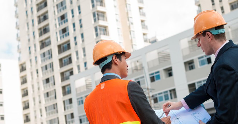 Dois homens estão em um canteiro de obra, olhando o prédio em construção e analisando os papéis